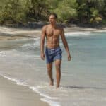 Confident man walking along a tropical beach in navy swim shorts, showcasing smooth, well-groomed skin under the sun.