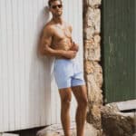 Shirtless man in light blue swim shorts leaning against a white wooden wall with a rustic green door and stone background, exuding confidence and a well-groomed appearance